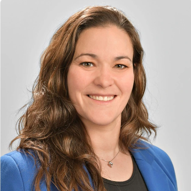 Woman with long brown hair wearing a blue blazer and black top, smiling at the camera against a plain light gray background.